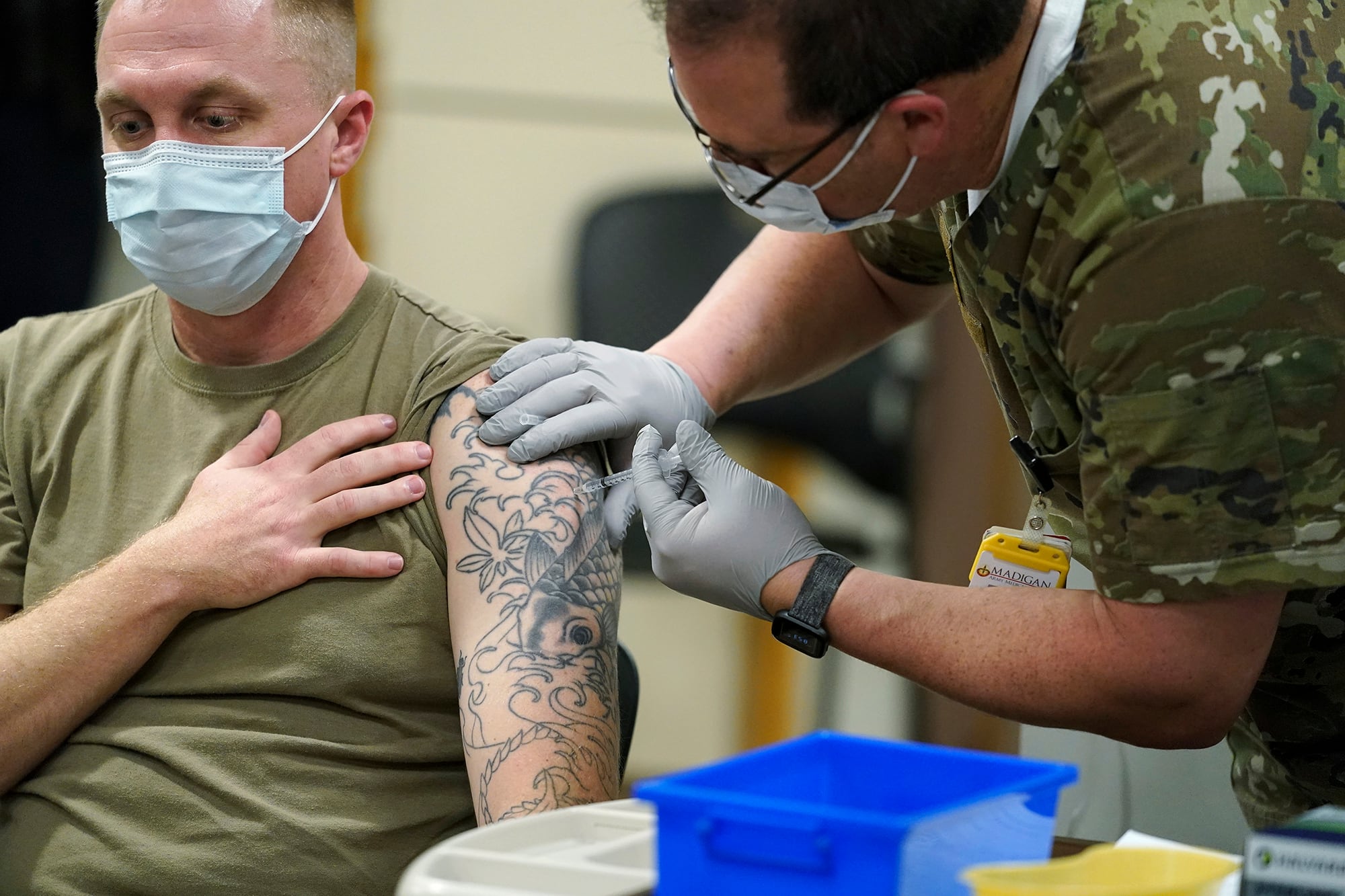 A seated and masked soldier receives a vaccination shot in his tattooed arm.