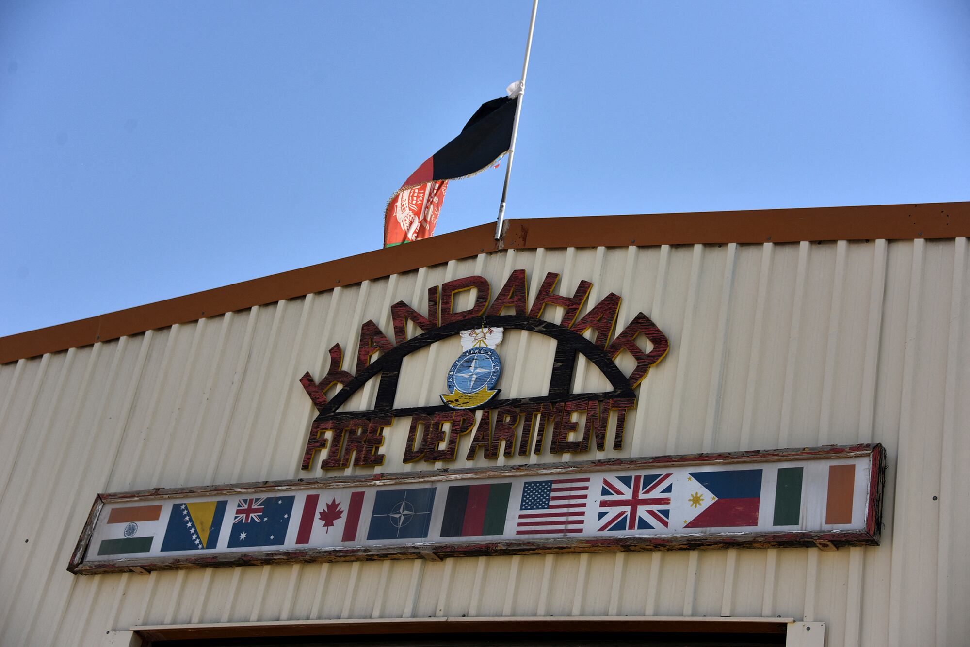 The Afghanistan flag flies over a sign that reads "Kandahar Fire Department" on the side of a building. The flags of NATO nations are displayed under the sign.