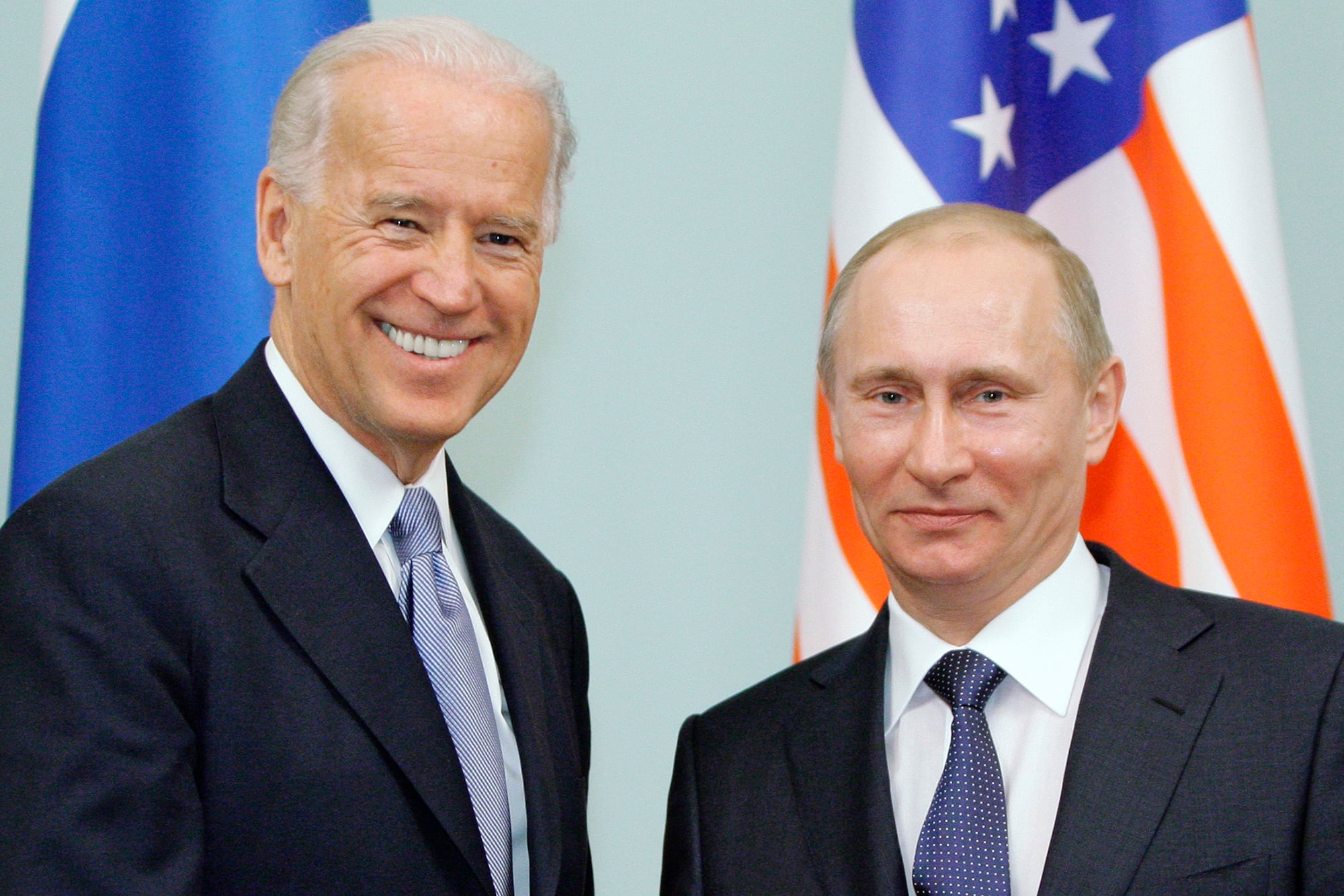 Joe Biden shakes hands with Vladimir Putin in front of flags.